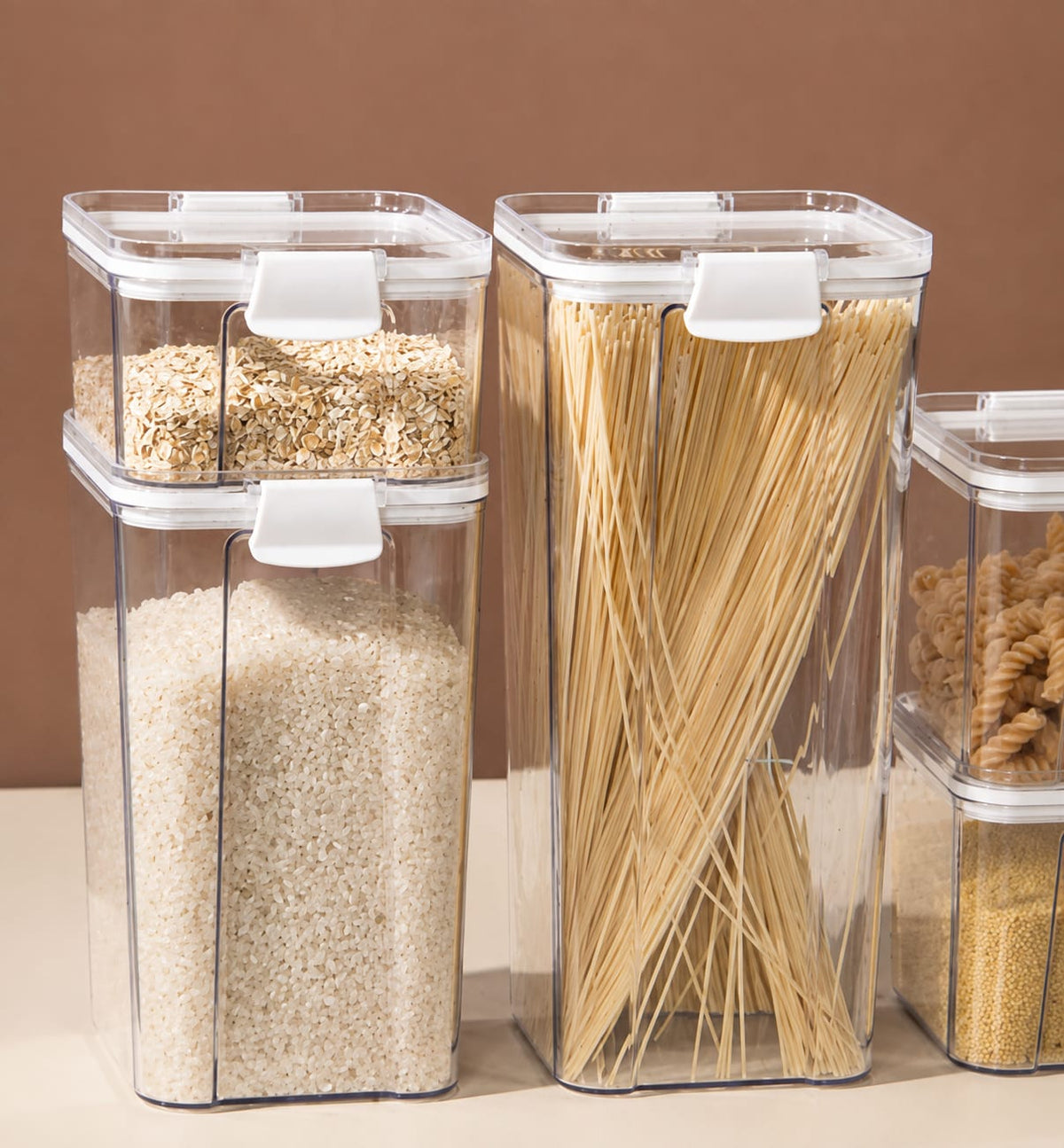 Clear plastic kitchen storage containers with white lids, filled with rice, pasta, and grains, arranged on a beige surface against a brown background.